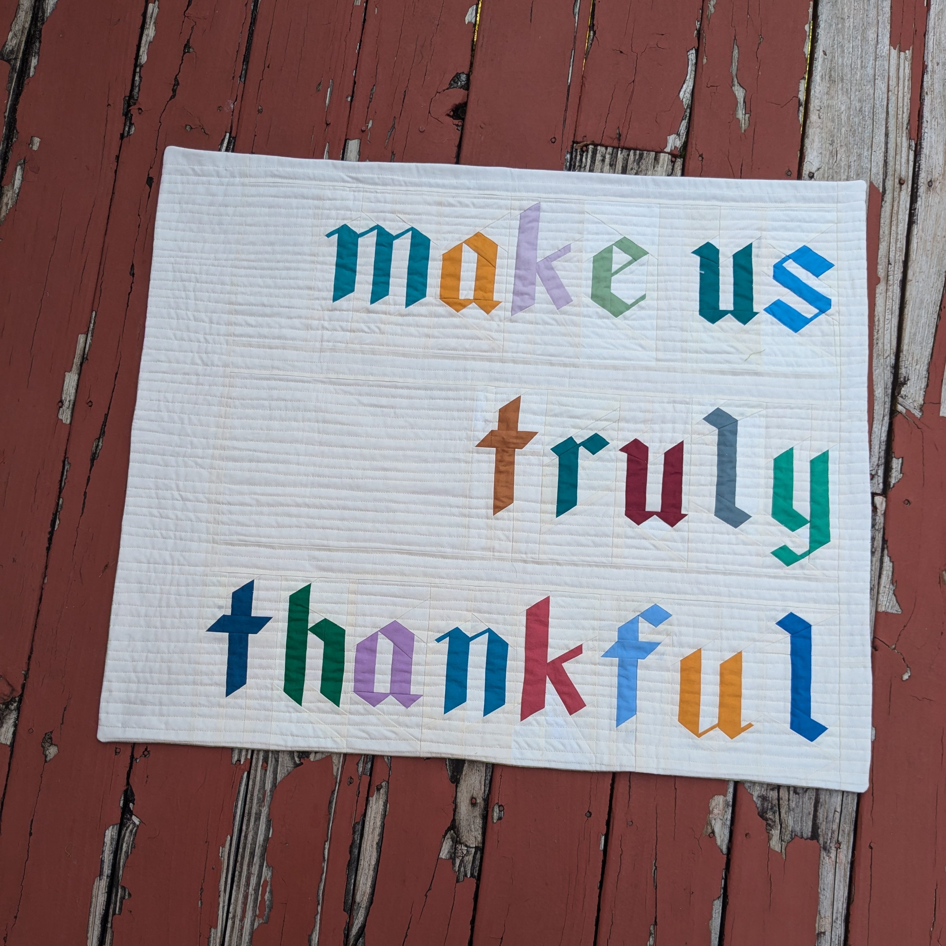 rectangular wall-hanging quilt with a white background and colored foundation pieced letters in a blackletter style. The letters read "make us truly thankful"